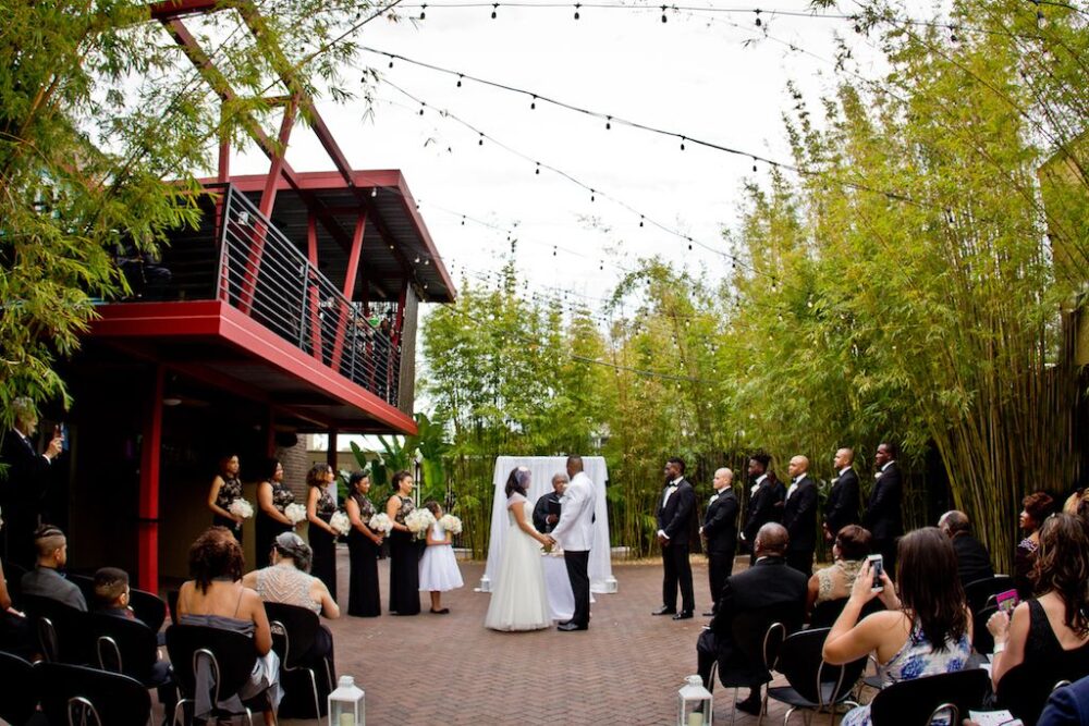 Elegant Black and White Outdoor St Pete Wedding Ceremony Portrait with Hurricane Lanterns on the Aisle, Bamboo and String Lights, and White Draped Ceremony Arch and Altar | Bride Wearing Cap Sleeve Ballgown Dress, Groom in White Jacket with Black Pants, Groomsmen in Black tuxedos, Bridesmaids in Black and Gold Dresses with White Bouquet | Garden Courtyard Ceremony at Unique Downtown St Pete Venue NOVA 535