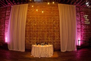 Elegant Black and White St Pete Wedding Reception Sweetheart Table with Textured Linen, White Draping, Hanging String Edison Lights on Exposed Brick Wall and Votive Candles | Unique Downtown St Pete Wedding Venue NOVA 535